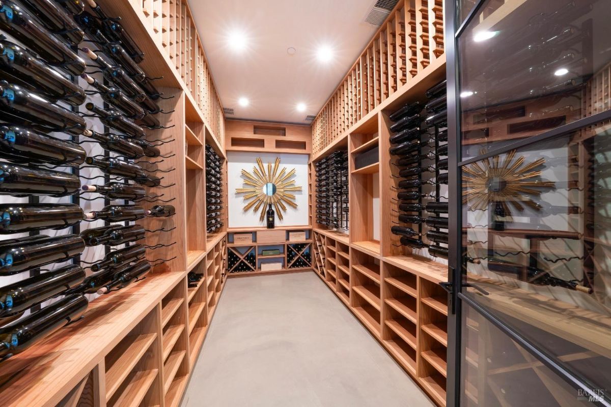 This wine cellar features wooden racks for storing bottles on both sides and compartments for additional wine storage below. A decorative sunburst mirror is mounted on the wall at the far end, adding a visual focal point to the space.