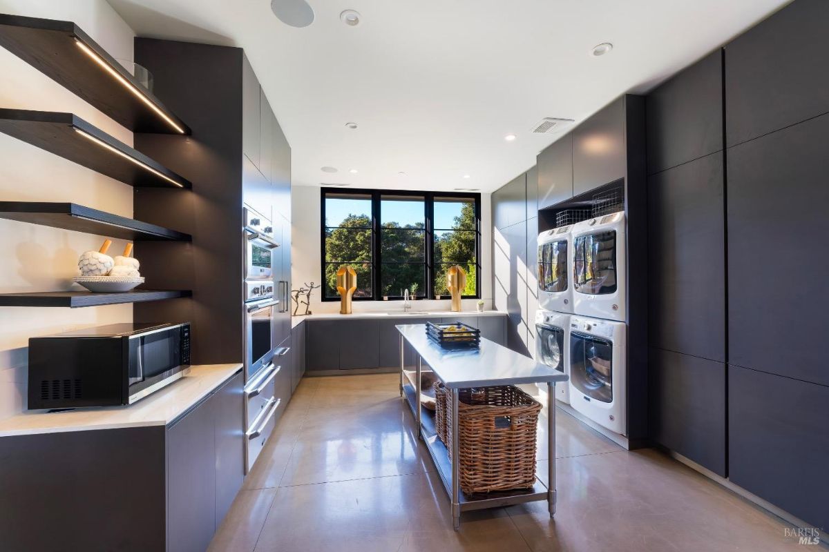 This room features a sleek design with dark cabinetry, built-in appliances, and open shelving. A central island with a wicker basket underneath complements the adjacent laundry machines and large windows overlooking greenery.