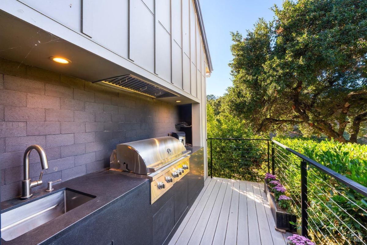 Outdoor kitchen features a built-in stainless steel grill with a black countertop and integrated sink. Adjacent deck has cable railings and overlooks lush greenery with planters lining the edge.