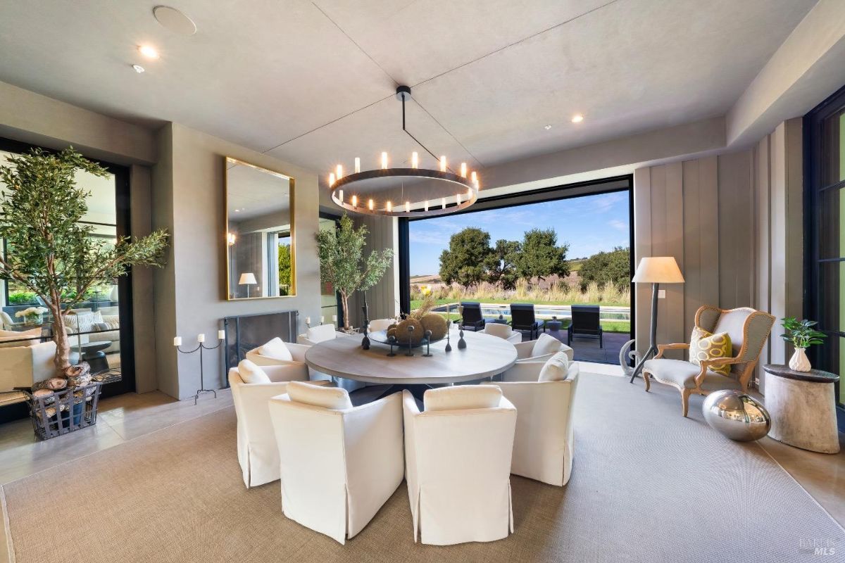 Dining area features a round table surrounded by upholstered white chairs and a large modern chandelier above. Sliding glass doors open to an outdoor seating area with expansive views of the surrounding landscape.