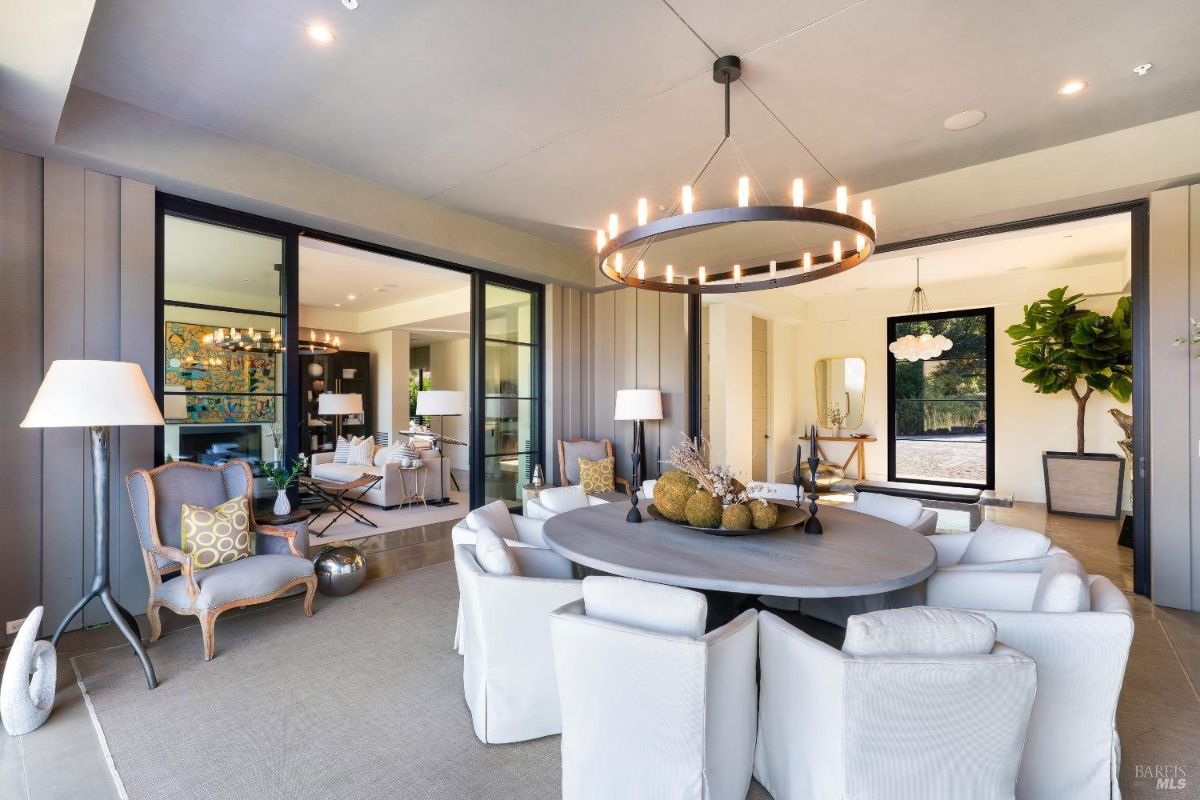 Dining space with a circular table surrounded by white upholstered chairs and a modern chandelier overhead. Open layout connects to adjacent living areas, featuring sleek furnishings and large windows for natural light.