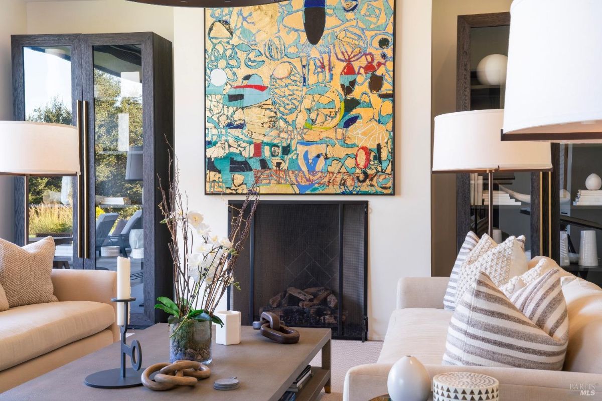 Seating area with neutral-toned sofas and a coffee table adorned with minimalist decor. Vibrant abstract artwork hangs above a black fireplace, flanked by sleek glass cabinets.