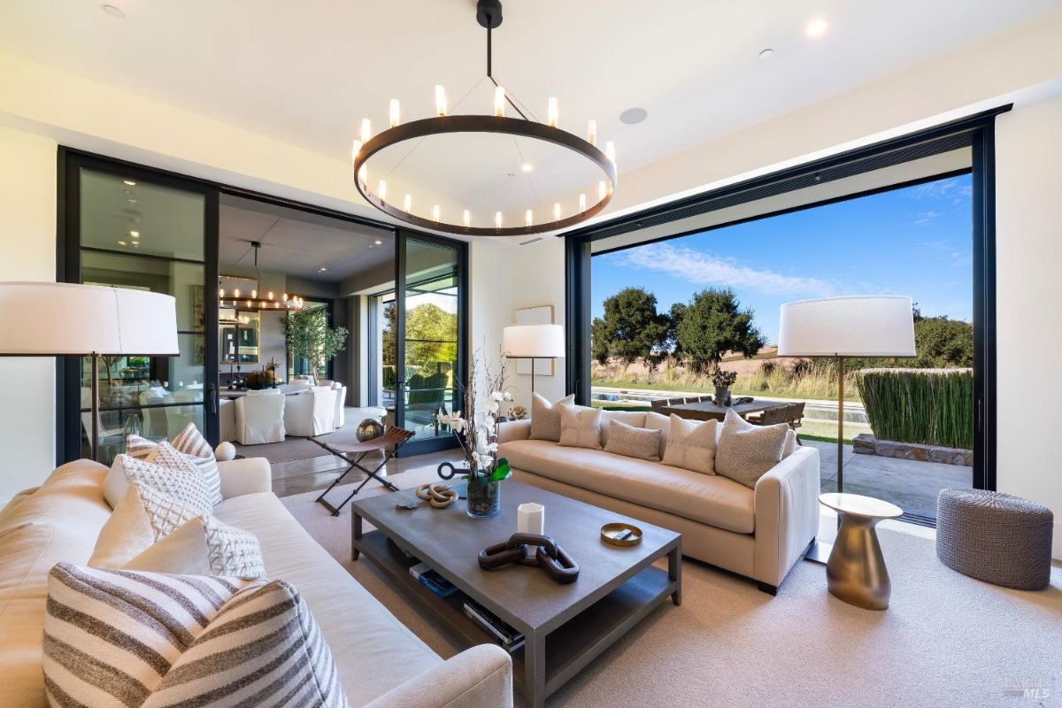 Modern living room with expansive glass doors opening to outdoor views and natural light. Comfortable seating arrangement with neutral tones and a central table enhances functionality and style.