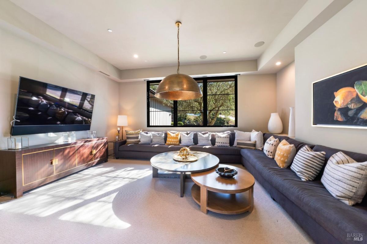 Living area with a large sectional sofa adorned with decorative pillows, centered around a modern round coffee table. Mounted television and a contemporary brass pendant light complete the space, which is illuminated by natural light from a large window.