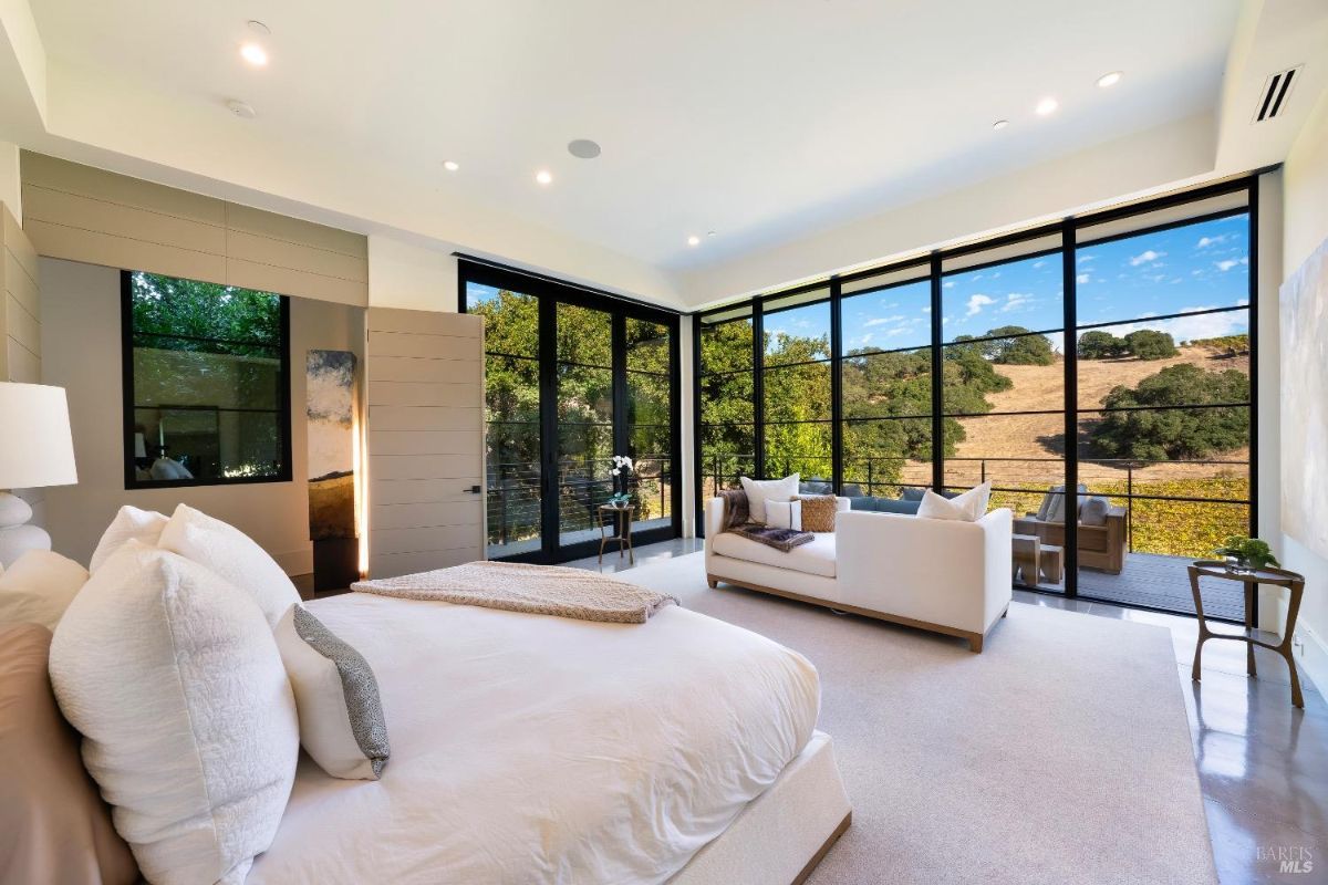 The bedroom features floor-to-ceiling windows offering expansive views of the surrounding hills and natural landscape. A cozy sitting area is arranged near the windows, complementing the minimalist yet luxurious interior design.