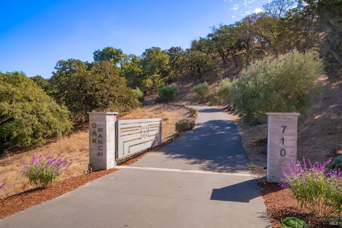 Entrance gate marked with "Oak Ranch" and address "710" is flanked by concrete pillars. Paved driveway winds uphill bordered by trees, shrubs, and lavender plants.