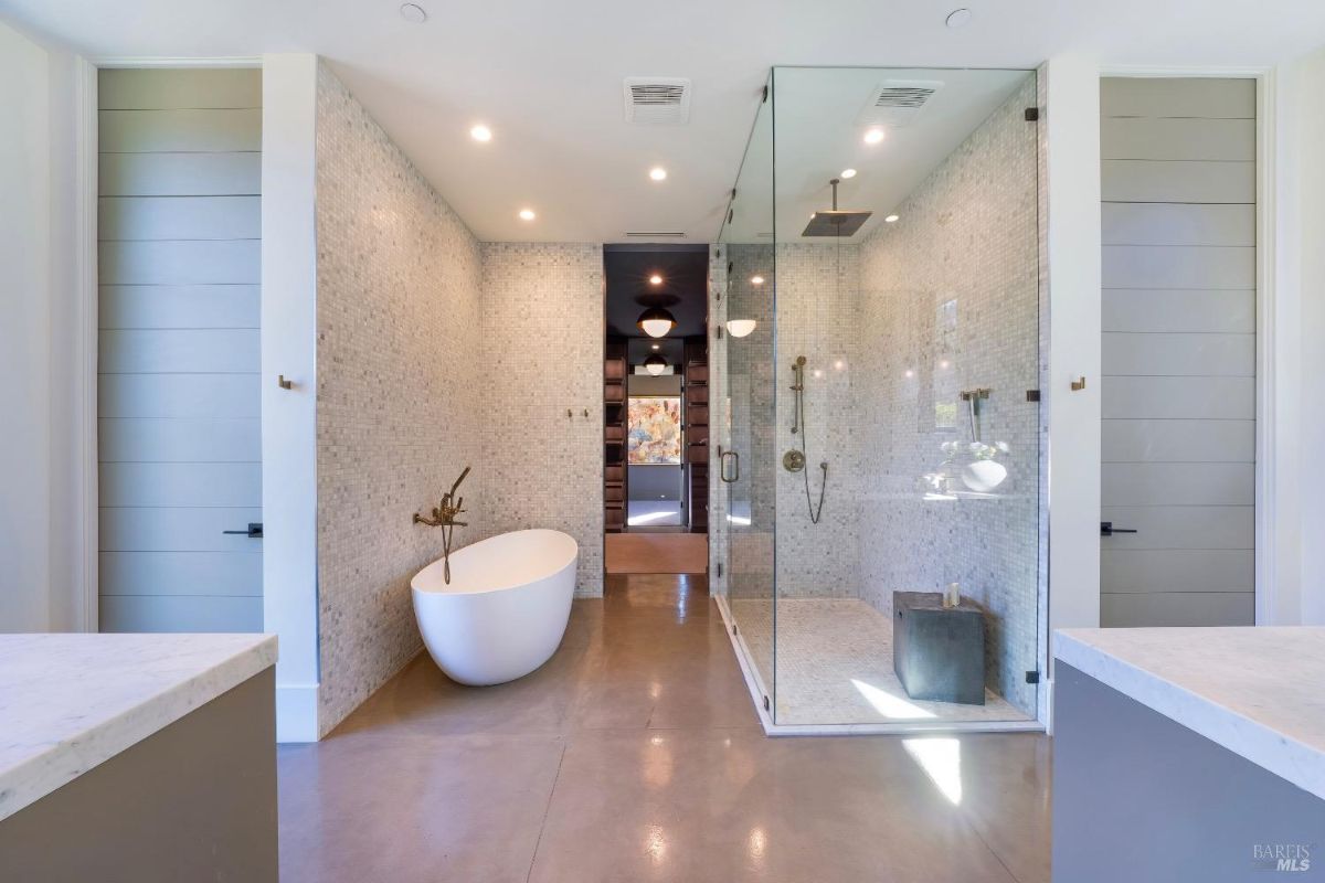 This bathroom features a freestanding tub with a wall-mounted faucet and a glass-enclosed walk-in shower with mosaic tile walls. Two doorways lead to adjacent areas, and the flooring is polished concrete.