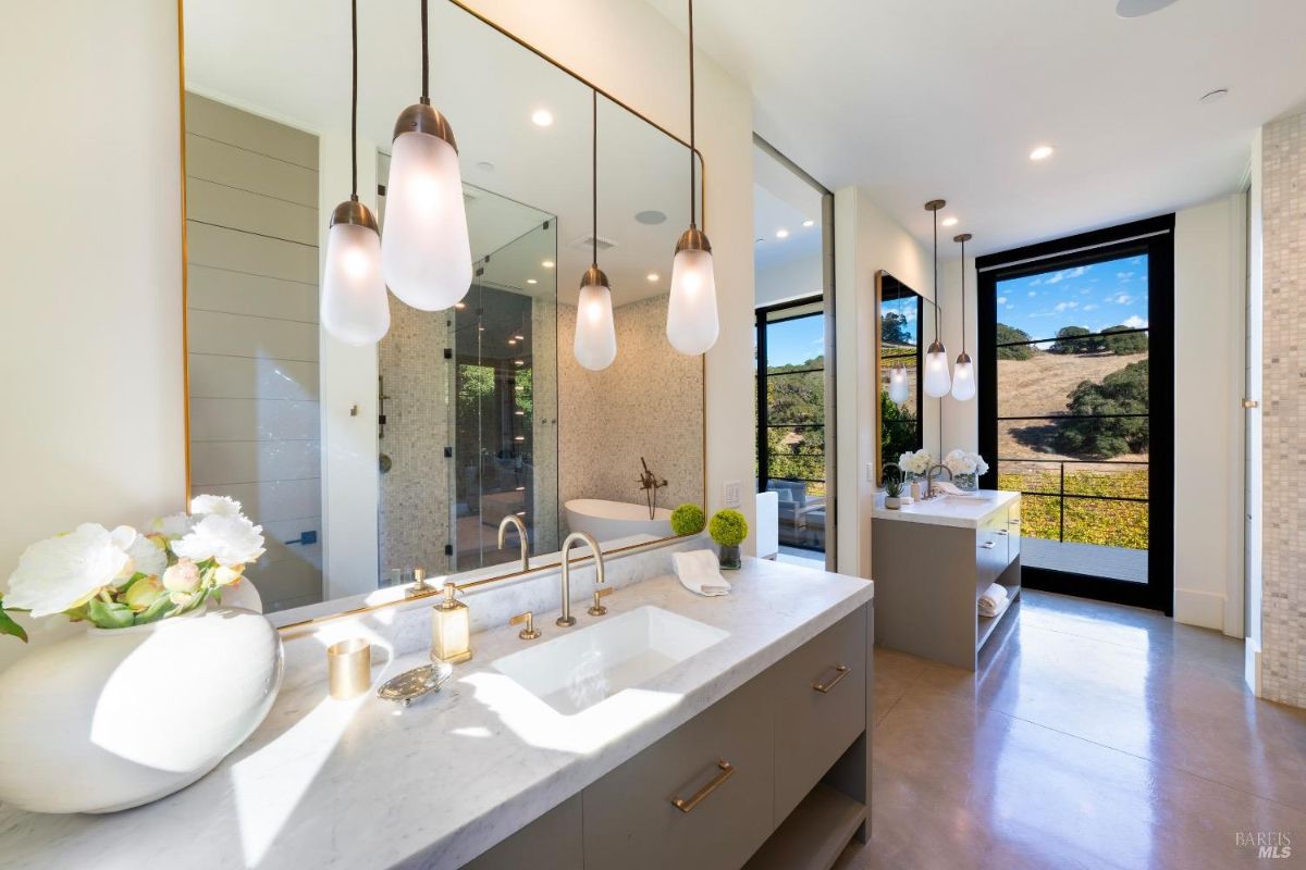 The bathroom features a marble countertop with dual sinks and brass fixtures, complemented by modern pendant lighting. Large windows provide natural light and offer views of the surrounding landscape, while a walk-in shower and freestanding tub are enclosed in a tiled area.