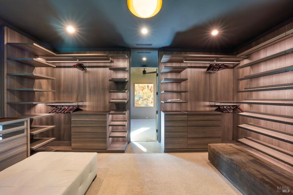 The walk-in closet features custom wood shelving and built-in drawers for organized storage. A bench and soft lighting create a functional and comfortable dressing area.