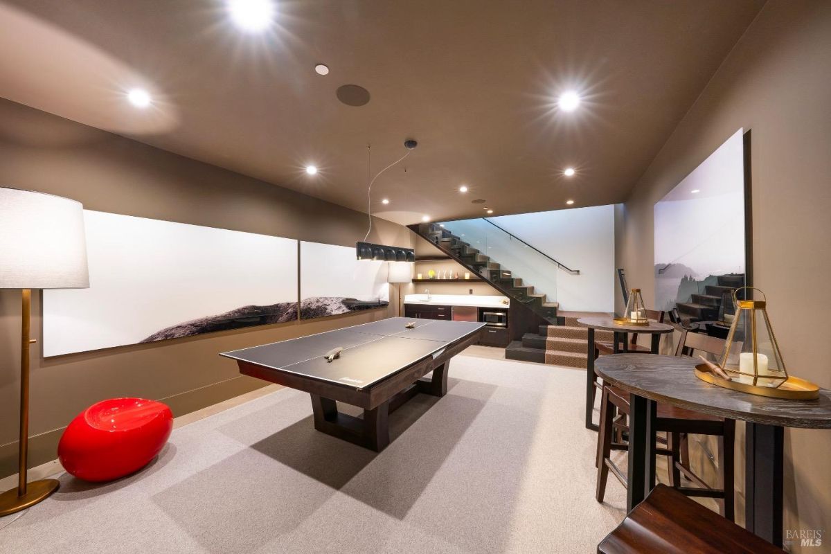 This room features a ping pong table as the centerpiece, surrounded by modern seating and lighting. A staircase with glass railings leads to an upper level, and a built-in bar area includes shelving and appliances.