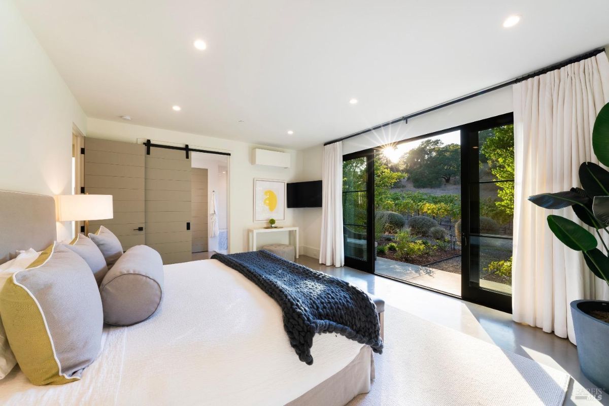 Bedroom features large sliding glass doors leading to an outdoor garden area. Interior includes neutral tones, a wall-mounted television, and a barn-style door separating the space from the en-suite bathroom.