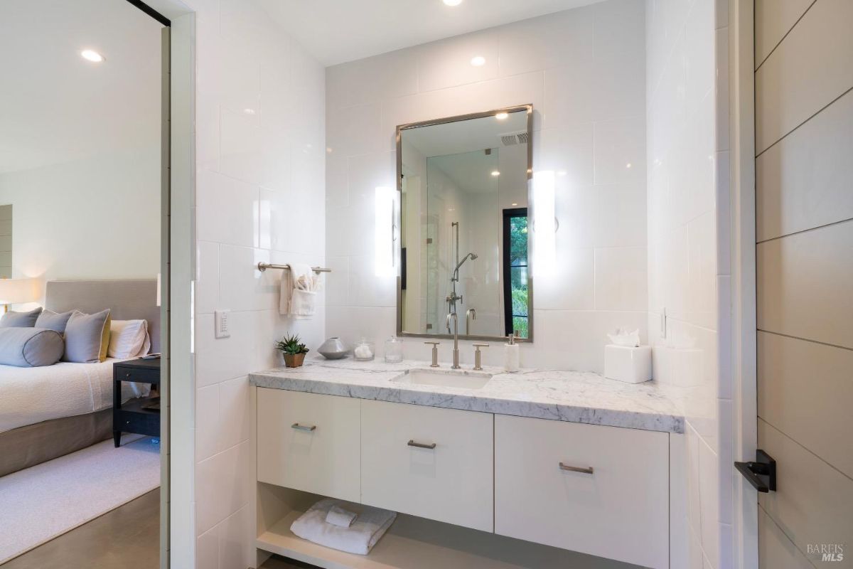 This bathroom features a marble countertop with a built-in sink and brushed metal fixtures. A large mirror with integrated lighting is mounted above the vanity, adjacent to a tiled wall and walk-in shower.