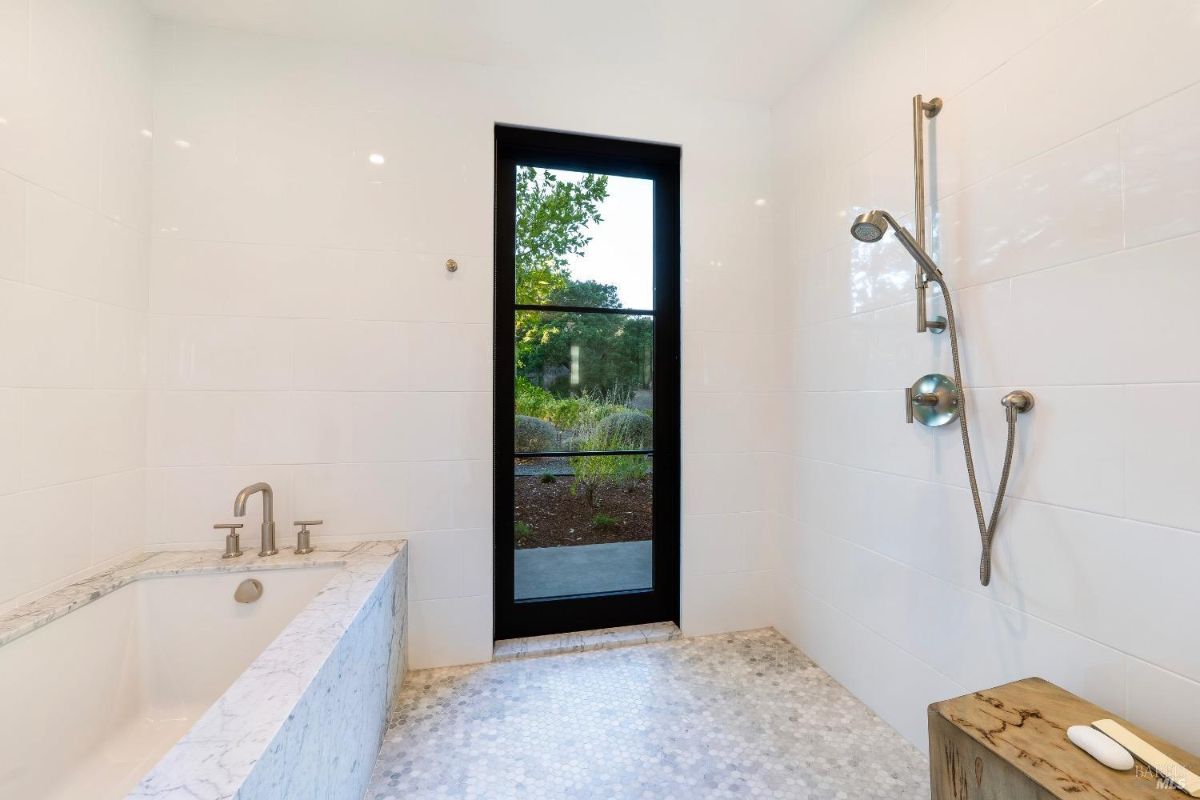 This bathroom features a marble-framed bathtub with brushed metal fixtures. A glass door opens to an outdoor view, complemented by a tiled shower area with a handheld showerhead.