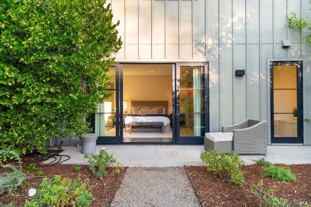 Exterior view shows a bedroom with sliding glass doors opening to a small patio area. Surrounding landscaping includes a large green tree, shrubs, and a gravel path leading to the entrance.