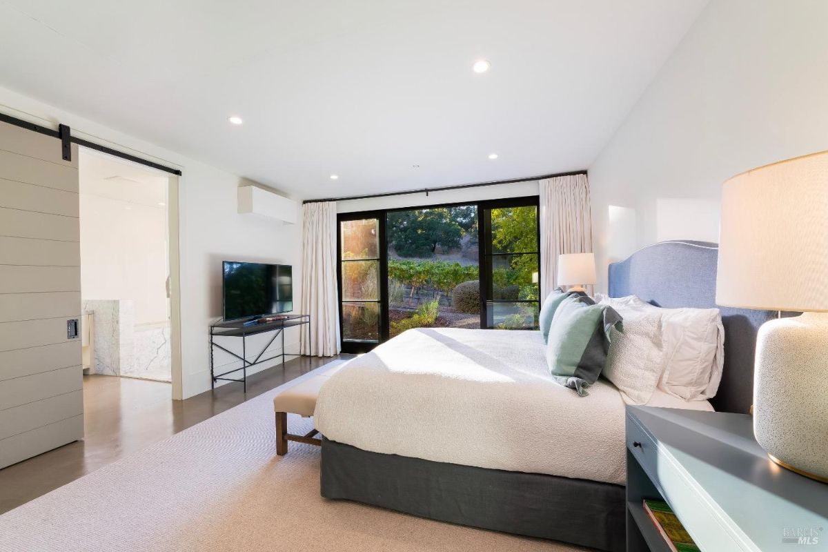 Bedroom features large sliding doors opening to a landscaped garden area with natural views. Interior includes a wall-mounted television, modern lighting, and an en-suite bathroom accessible through a barn-style door.