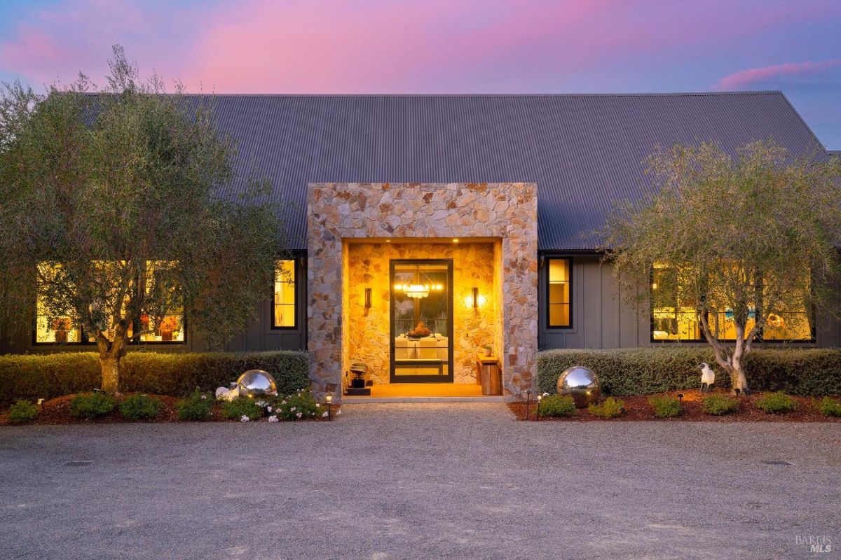 Stone-clad central entrance features a glass door framed by warm lighting. Flanking windows and neatly trimmed shrubs accentuate the structure, set under a large sloped roof.