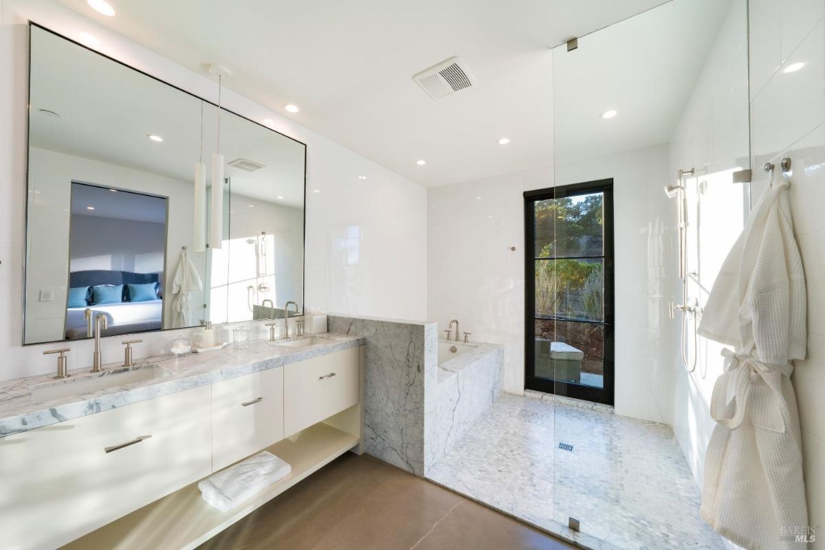 This bathroom features a double sink vanity with a marble countertop and a large wall mirror. A walk-in shower and a marble-framed bathtub are located near a glass door that opens to an outdoor view.