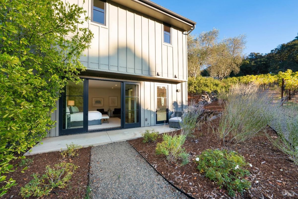 Two-story building features sliding glass doors on the ground level, opening into a bedroom. Gravel path leads through landscaped shrubs and mulch, with vineyards visible in the background.