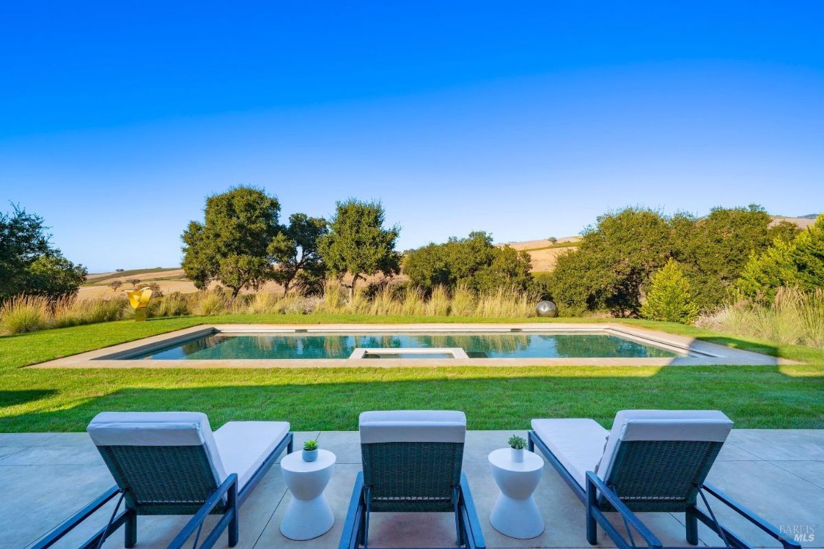 Rectangular pool with an integrated hot tub is surrounded by manicured lawn and natural grasses. Chaise lounges with side tables are positioned on a paved area overlooking the pool and surrounding landscape.