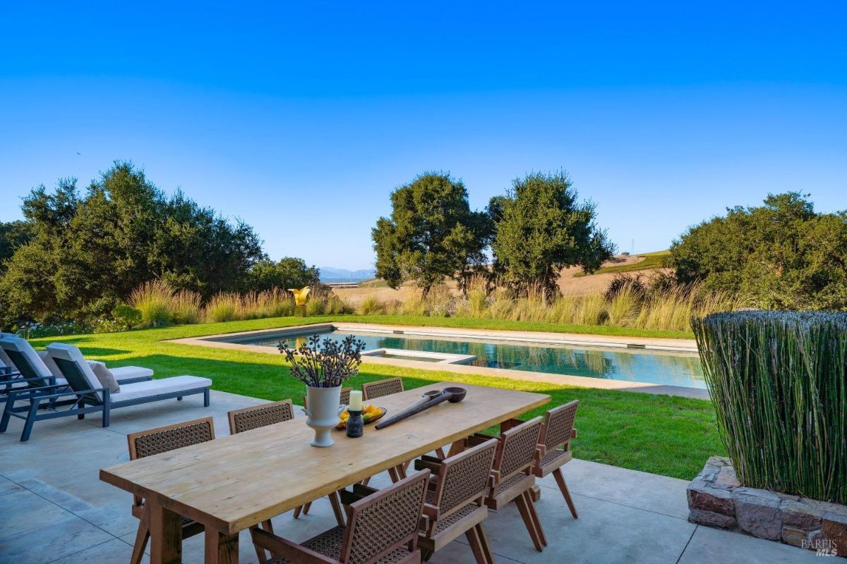 Outdoor wooden dining table with woven chairs is placed on a concrete patio near a grassy lawn. Rectangular pool and lounge chairs are set against a backdrop of trees and rolling hills.