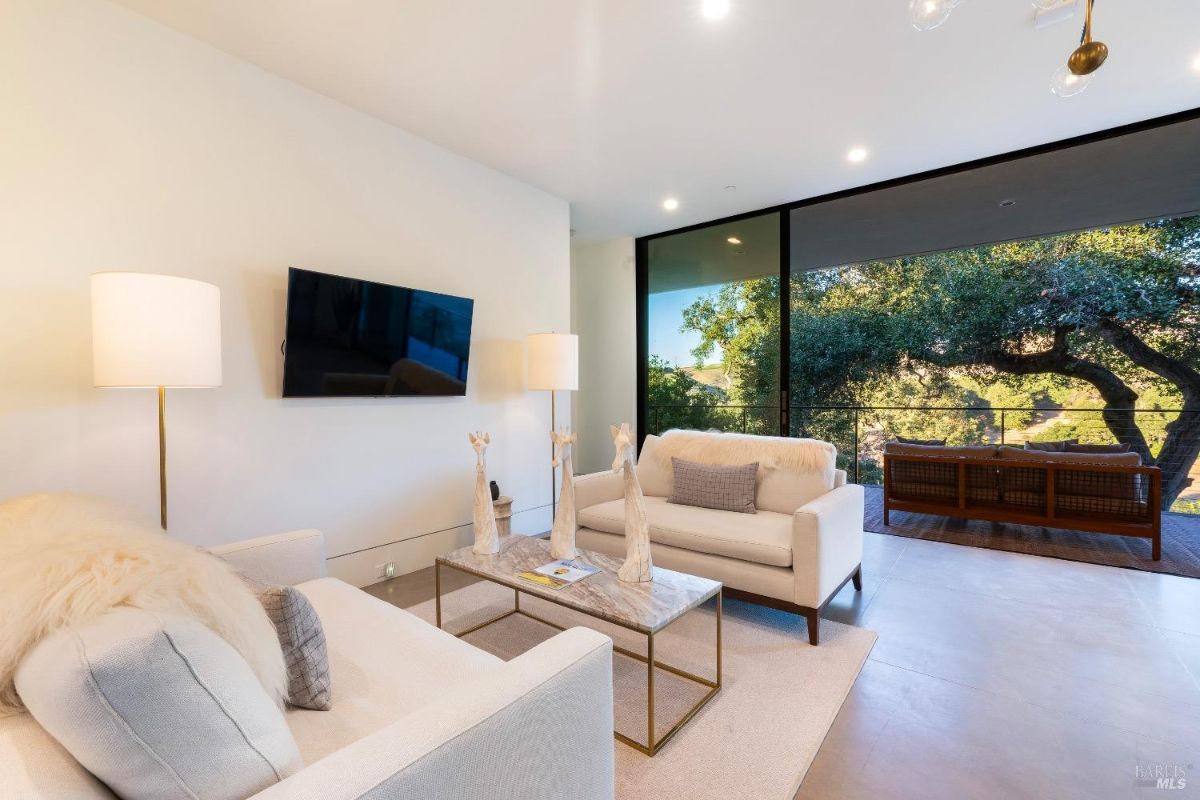 Living area with minimalist design includes neutral-toned furniture and a mounted television. Floor-to-ceiling glass doors open to a balcony with views of surrounding trees, providing a connection to nature.