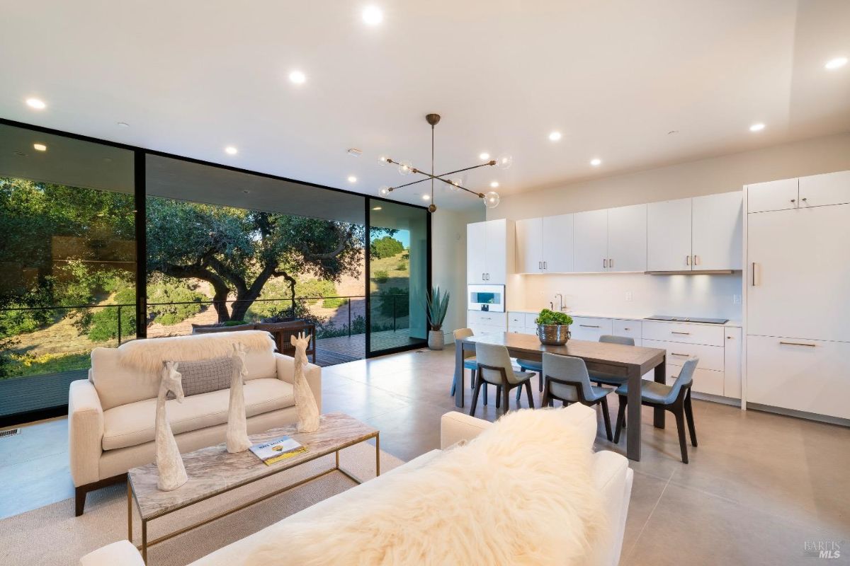 Open-concept living and dining area with modern furnishings and minimalist decor. Floor-to-ceiling glass doors provide natural light and views of the surrounding landscape.