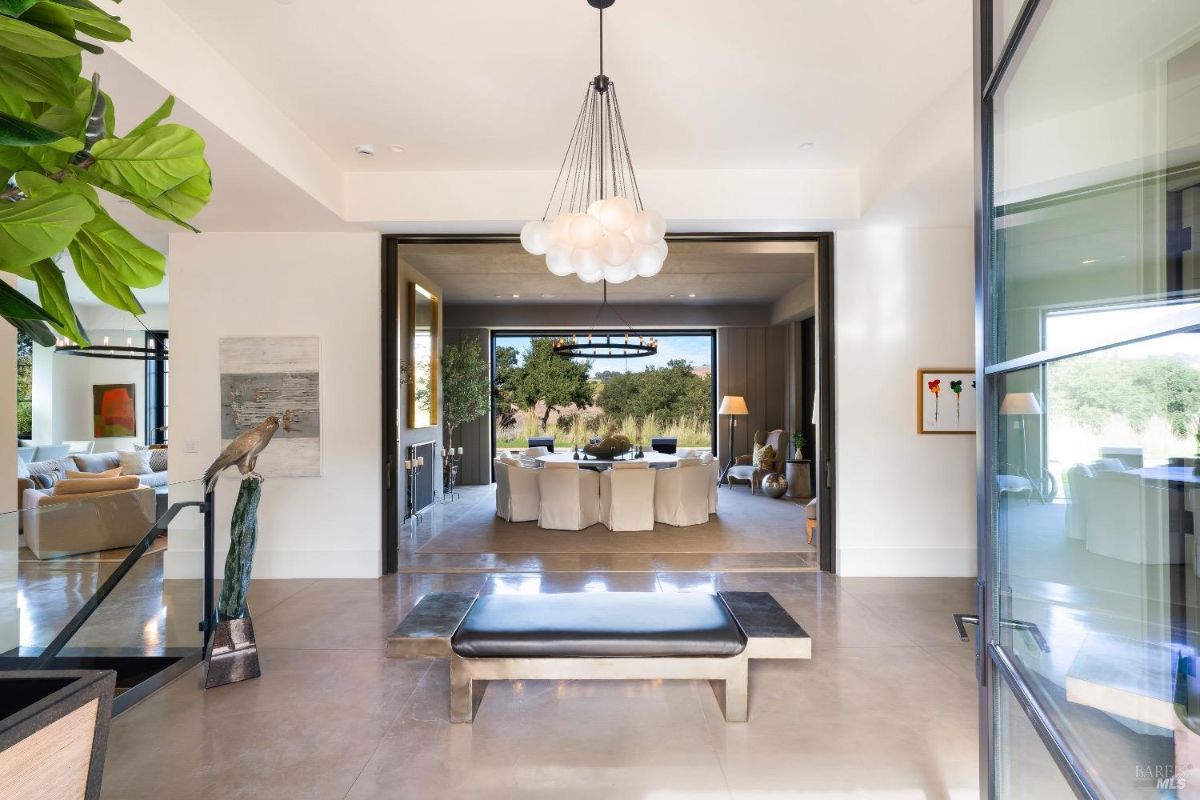 Spacious entryway featuring polished floors, a modern bench, and a central sculptural chandelier. Open layout leads to a dining area with large windows showcasing outdoor views.