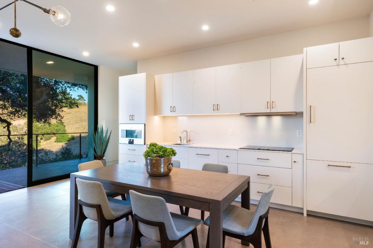 Minimalist kitchen design with white cabinetry and integrated appliances. Adjacent dining area includes a modern wooden table and upholstered chairs, complemented by large glass doors with outdoor views.