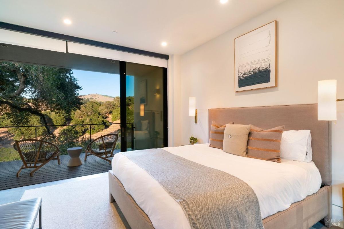 Bedroom features floor-to-ceiling glass doors opening to a private balcony with views of rolling hills. Neutral decor with modern accents creates a calming and elegant space.