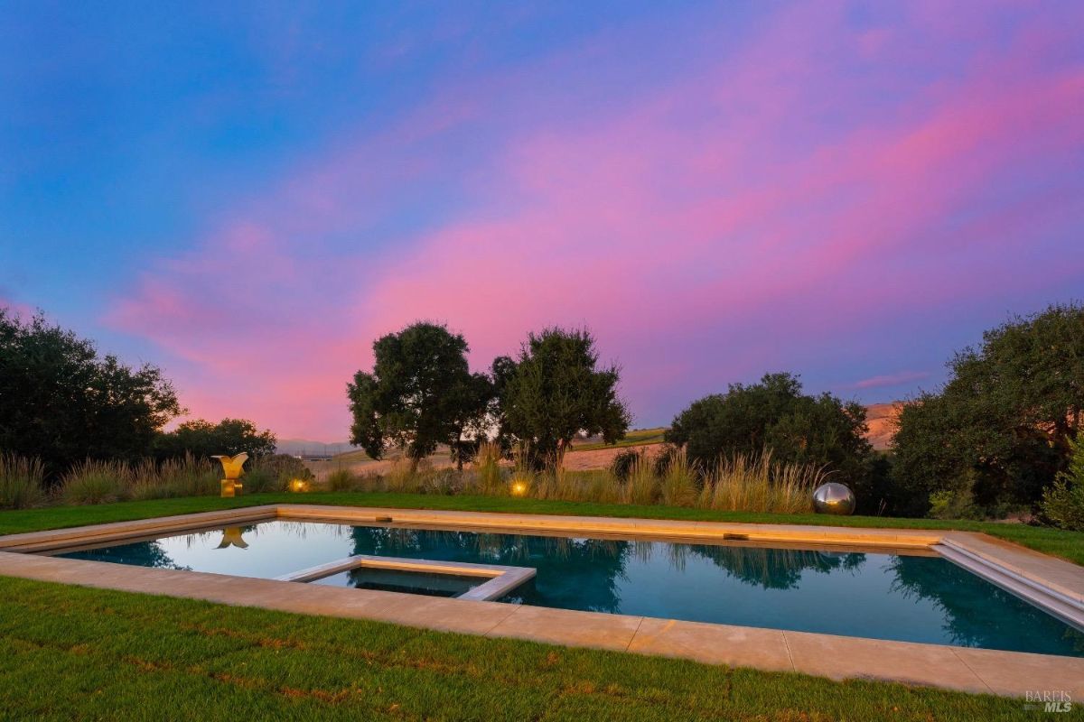 Rectangular pool with integrated hot tub is surrounded by a manicured lawn and decorative grasses. Trees and open fields create a natural backdrop under a colorful sunset sky.