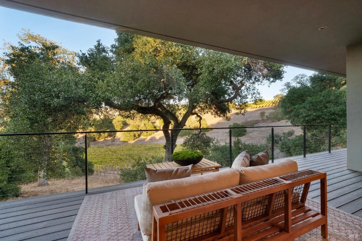 A wooden outdoor sofa with beige cushions is placed on a rug on a deck. A glass railing provides views of trees and rolling hills with vineyards in the distance.