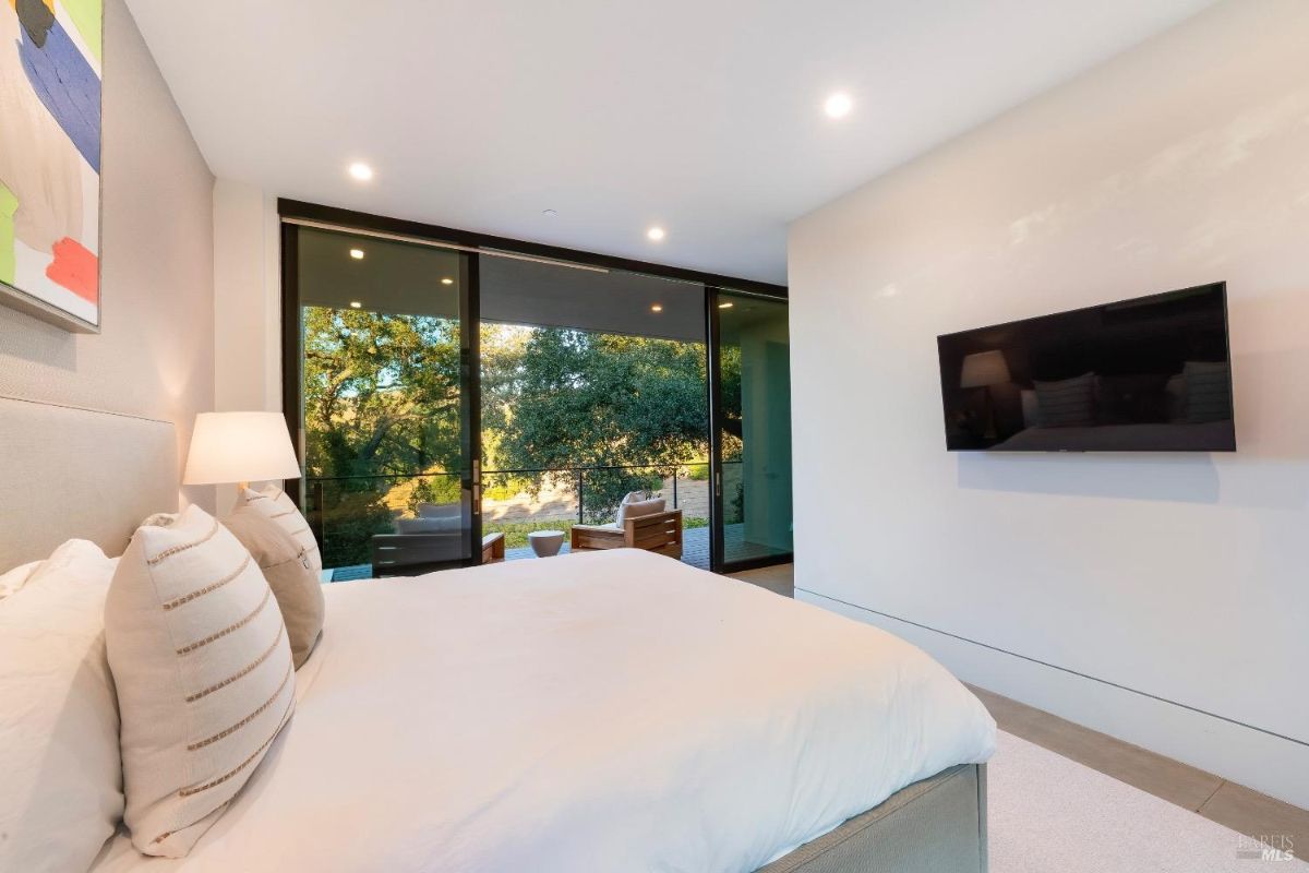 Bedroom includes floor-to-ceiling sliding glass doors that open to a private patio with outdoor seating. Wall-mounted television complements the minimalist design and neutral decor.