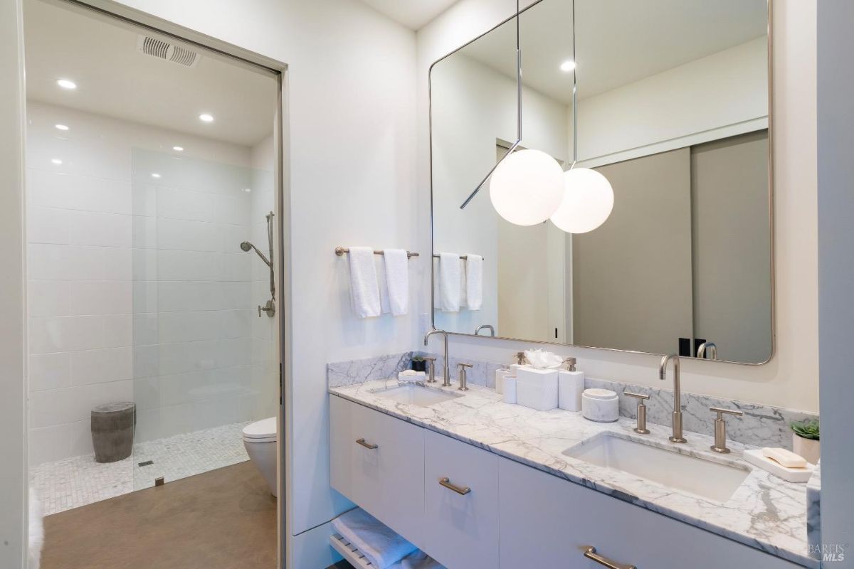 This bathroom features a marble countertop with dual sinks and brass fixtures beneath a large rectangular mirror. A walk-in shower with white tile and a glass enclosure is located adjacent to the vanity area.