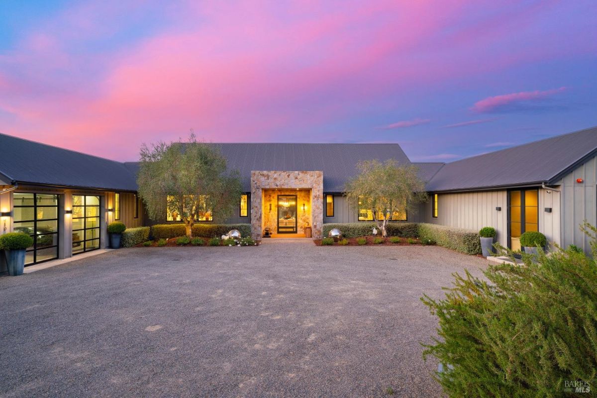 Single-story home with a central stone entrance flanked by large windows. Landscaped with shrubs, small trees, and potted plants, set against a gravel driveway and illuminated under a colorful evening sky.