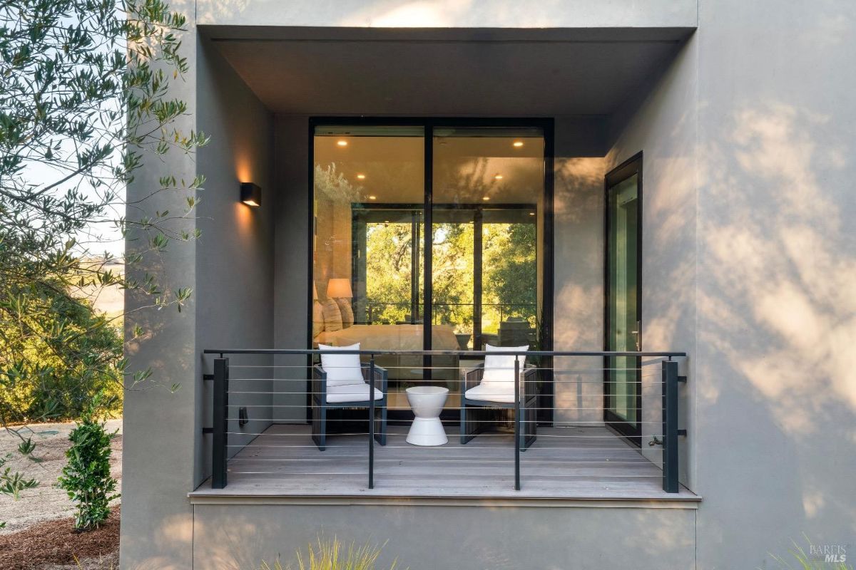 This patio features two white cushioned chairs with a small white table in the center, enclosed by a black railing. Large glass sliding doors connect the patio to an interior room, allowing for an open view of the surrounding greenery.