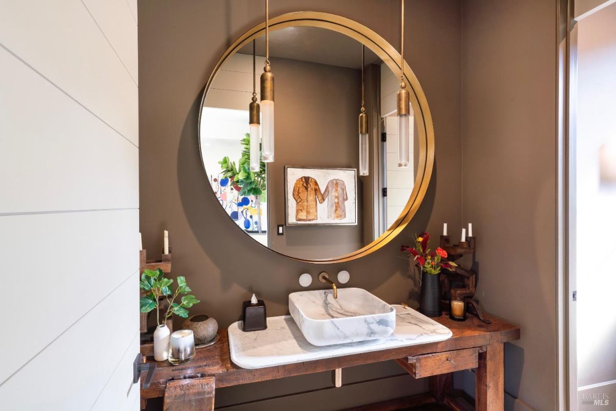 The bathroom features a large circular mirror framed in gold, accompanied by modern pendant lighting. A marble sink rests on a rustic wooden countertop, blending contemporary and natural design elements.