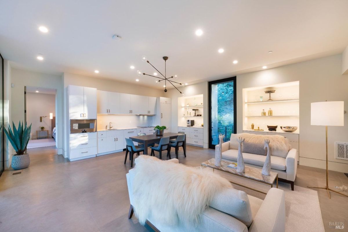 Open-concept living space with a modern kitchen featuring sleek white cabinetry and integrated appliances. Cozy seating area and dining table are complemented by built-in shelving and a large window providing natural light.