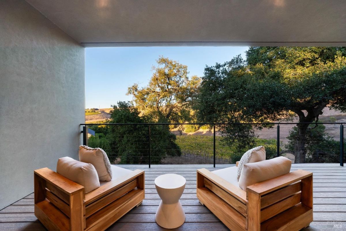 Two wooden lounge chairs with white cushions are placed on a deck with a small white table in between. A railing frames the space, offering a view of trees and a sloping hillside in the distance.