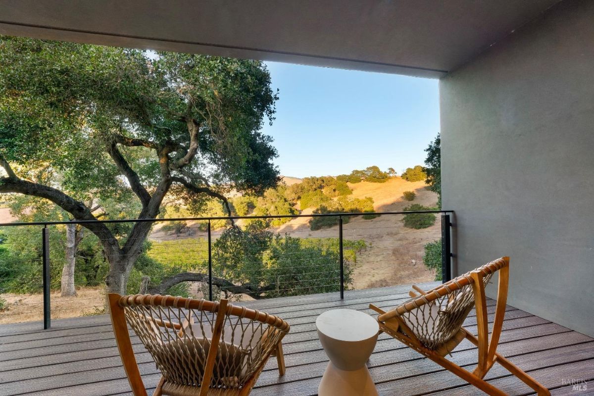 This balcony features two wooden lounge chairs with a small white table between them. A railing encloses the space, offering an unobstructed view of trees and rolling hills.