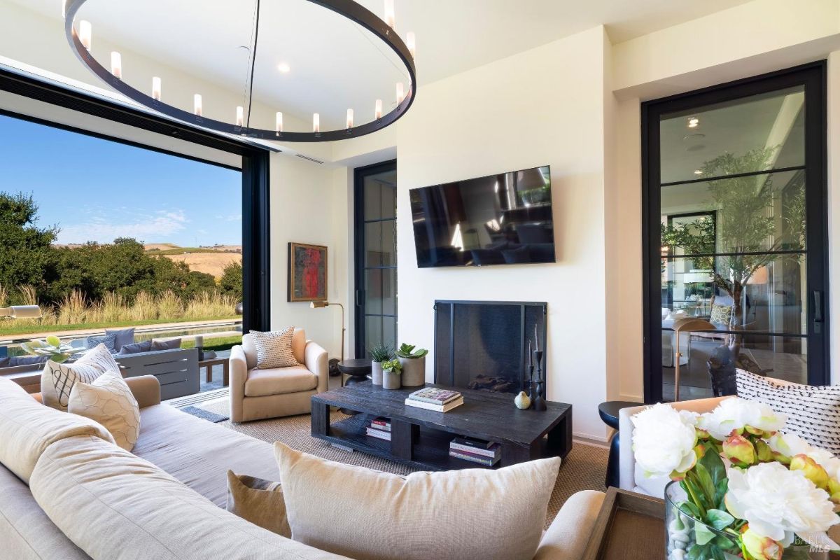 Living room with neutral-toned furniture and a mounted television above a fireplace. Glass doors open to an outdoor seating area with views of the surrounding landscape.