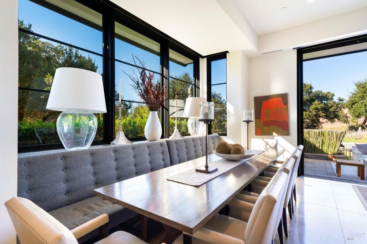 Dining area with a modern table and upholstered bench seating positioned near large windows. Natural light highlights contemporary decor and views of the surrounding greenery.