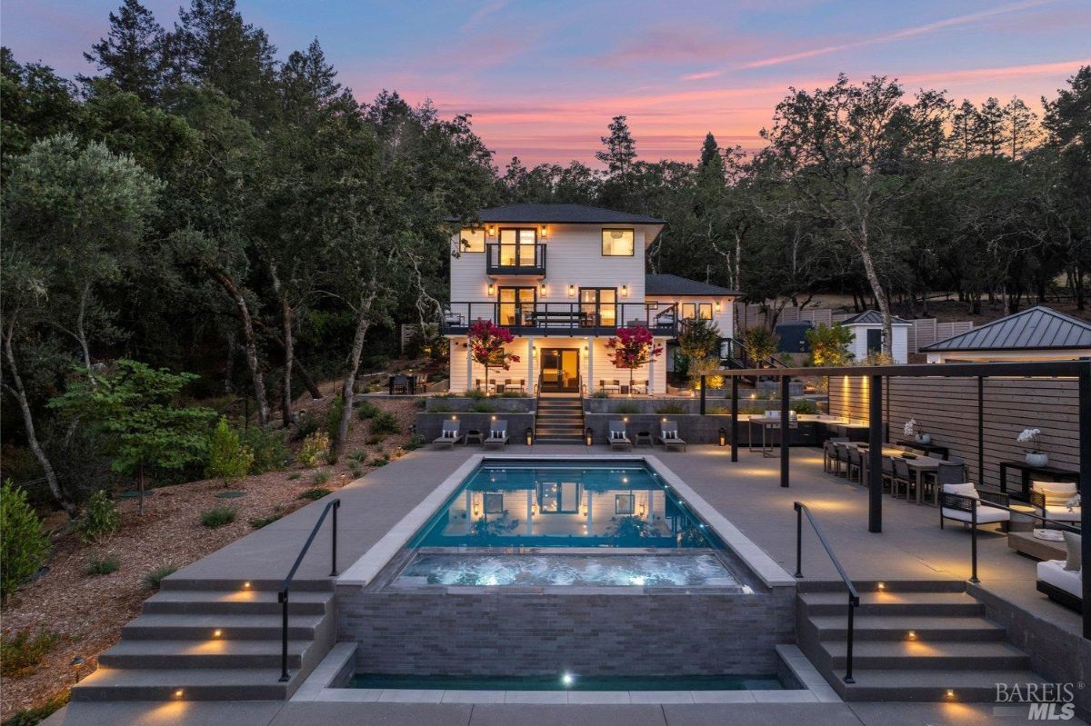 A modern, multi-story home with a beautiful outdoor pool and hot tub, surrounded by a lush forested area, lit warmly at dusk.