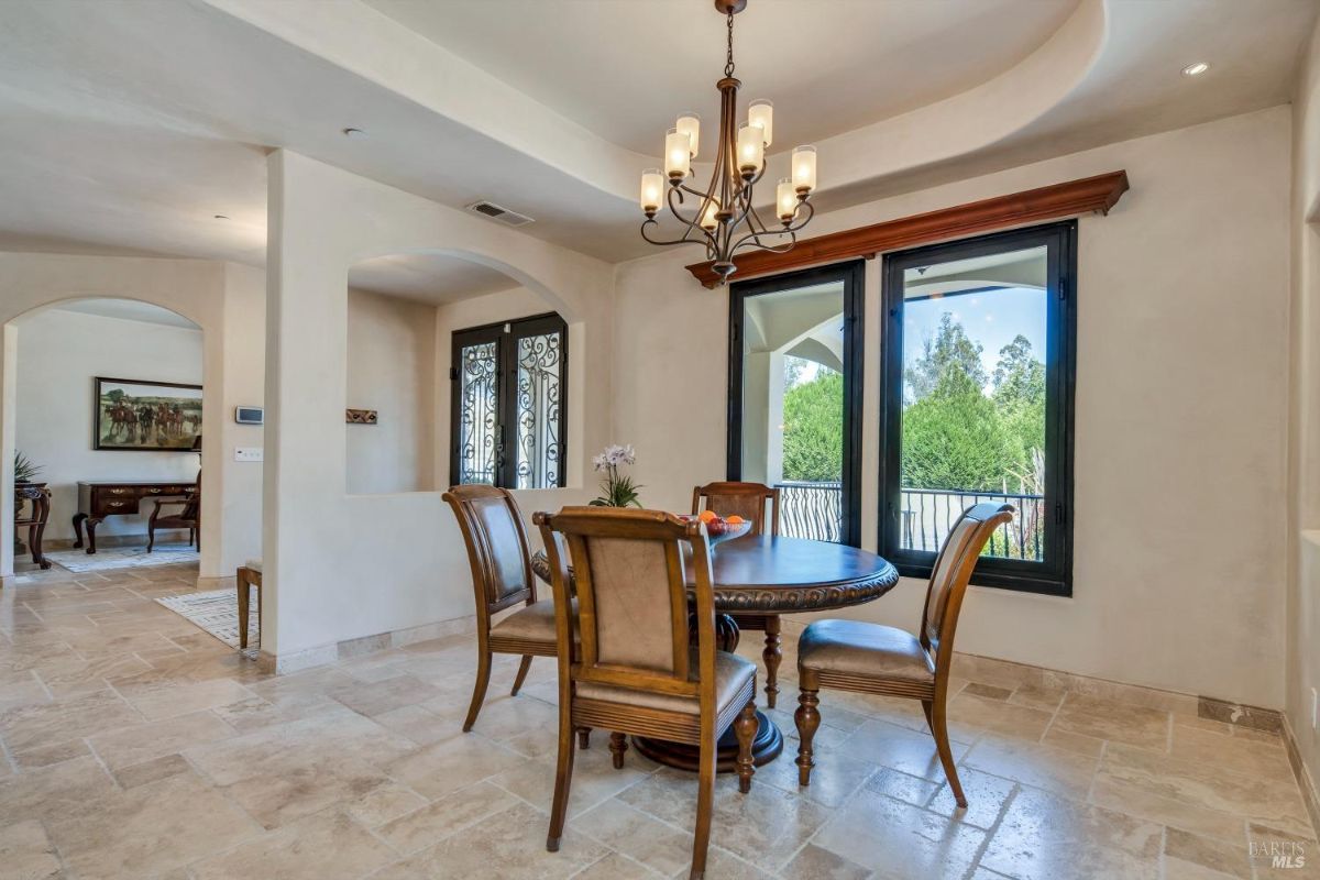 A round dining table with four chairs, a chandelier, and a view of a balcony and garden through large windows.