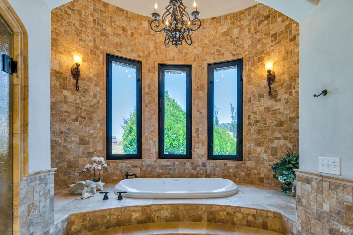 The bathroom features a large bathtub, three windows, and a chandelier.