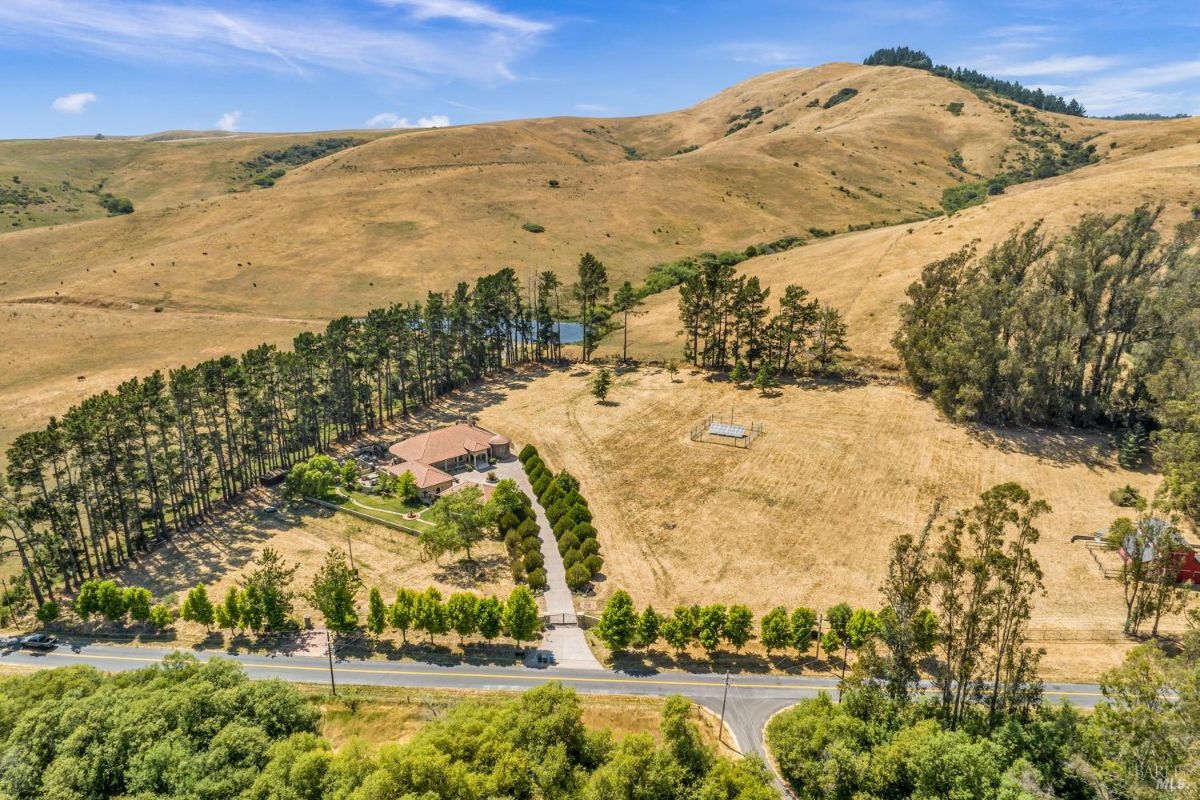 Wide-angle view of the property showing the house, a tree-lined driveway, and surrounding hills.