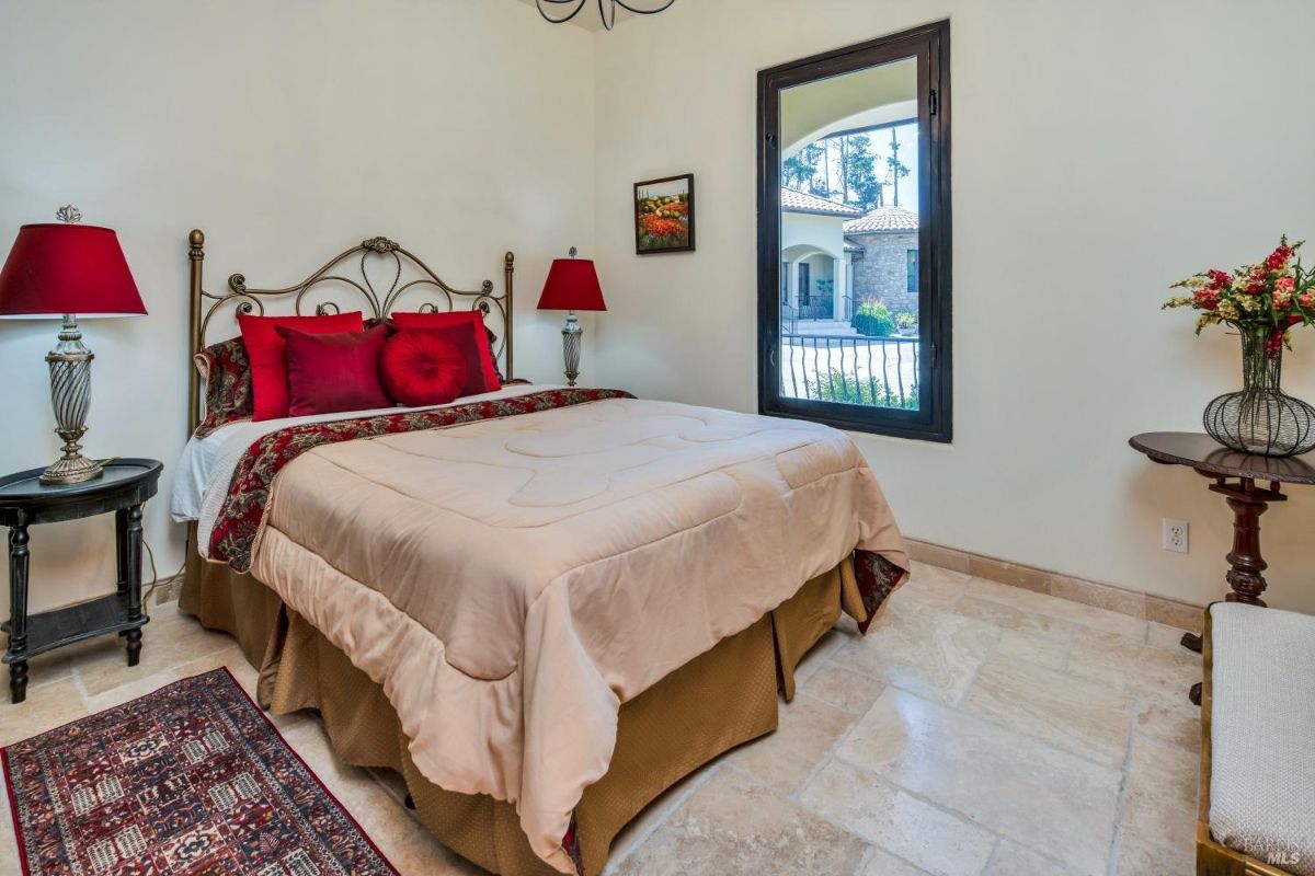 The bedroom features a bed with a beige comforter, red pillows, and a view of the outdoors.