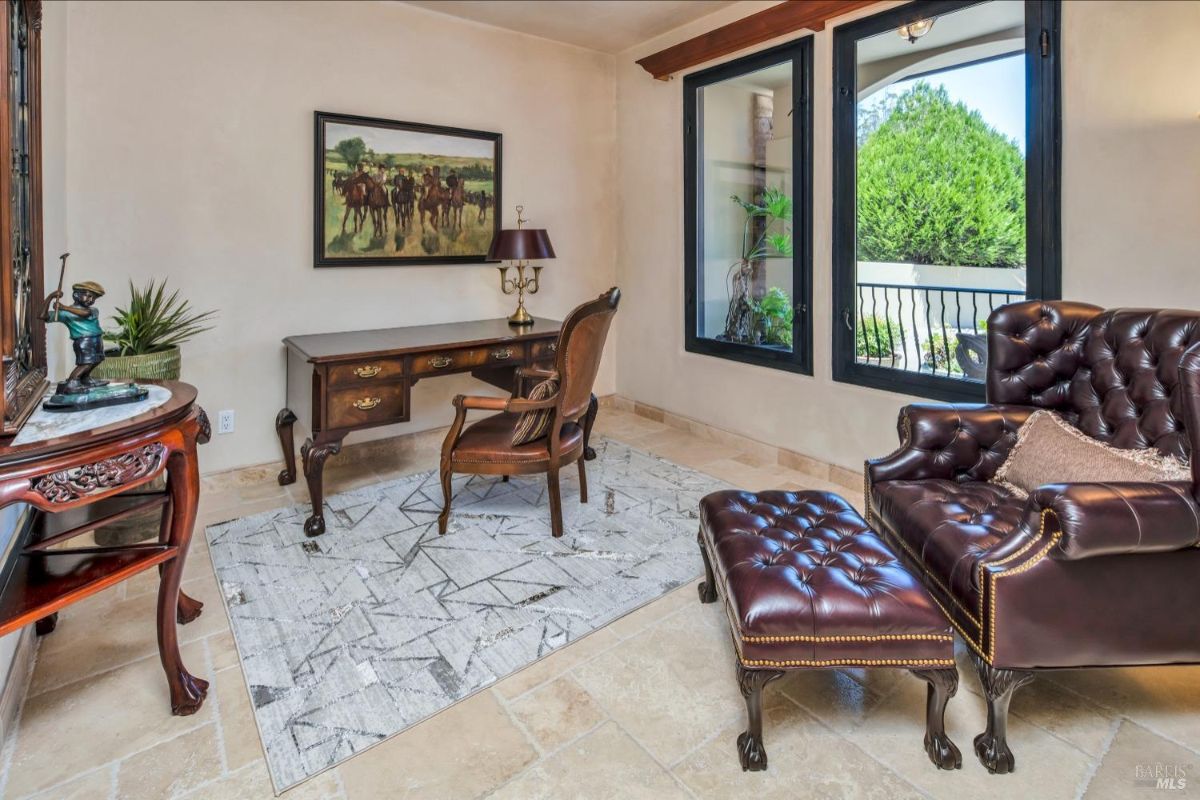 Office space with a classic wooden desk, leather armchair, and large windows overlooking greenery.