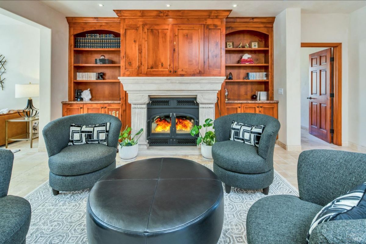 Sitting area with a fireplace, built-in wooden shelves, and gray chairs surrounding a round ottoman.