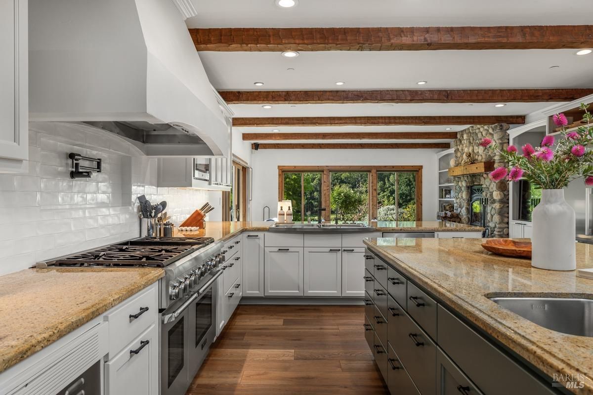 This kitchen emphasizes natural light with large windows overlooking greenery. White cabinetry, rustic wooden beams, and a combination of modern appliances make it a functional yet cozy space.