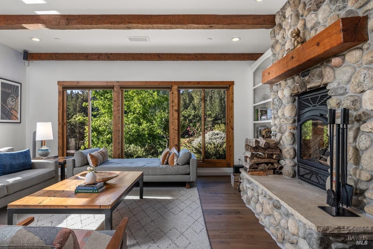 A cozy living room with a stone fireplace, large windows, and a comfortable seating arrangement. The room has a rustic charm with exposed wooden beams and a wood-burning fireplace.